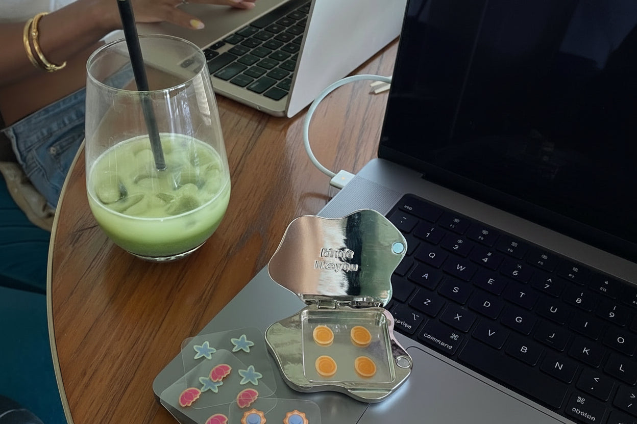 A styled desk scene showing a person using a laptop next to a green drink in a glass, with a small clear cosmetic case containing orange circular items and decorative flower stickers on a wooden table.