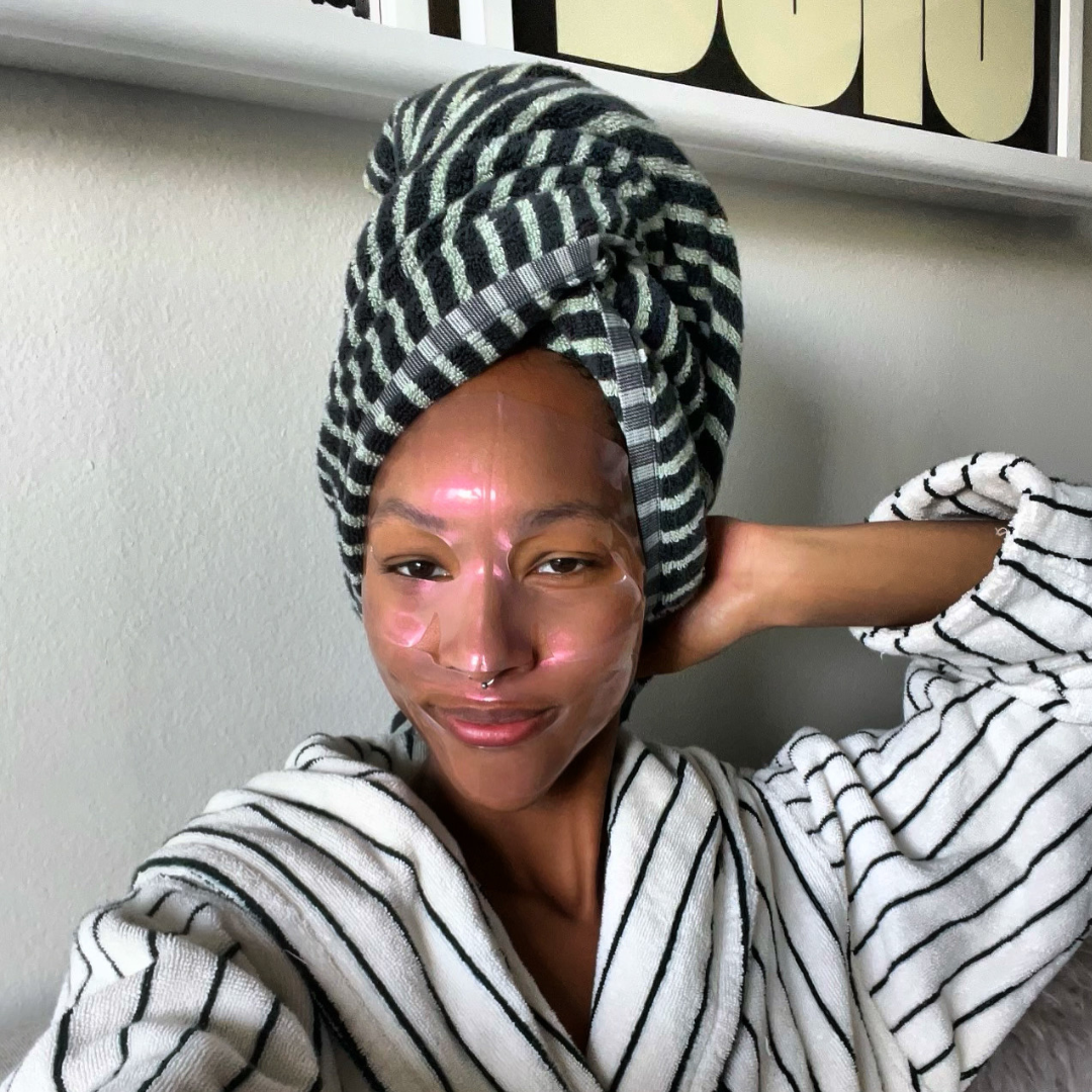 Woman wrapped in a striped towel with a neutral background wearing a dewy mask called aura drip